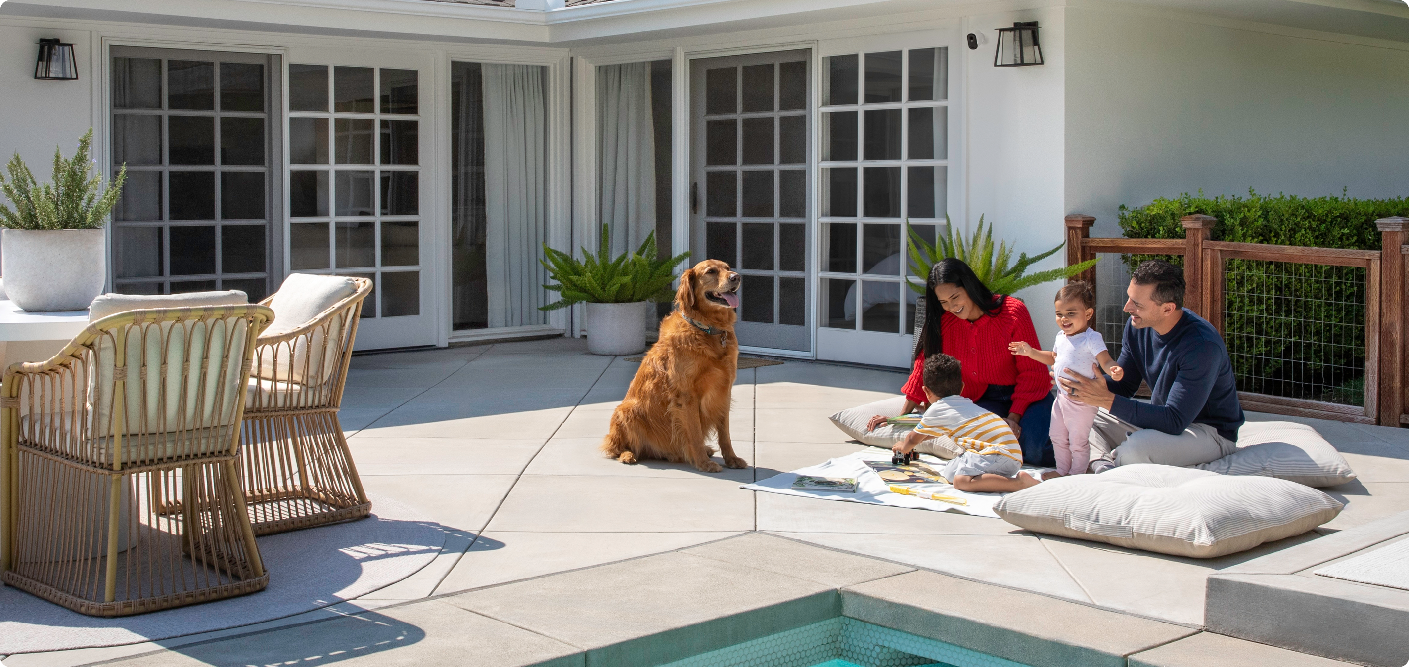 A dog with a mom and child outside by a pool with the Arlo Essential 3rd Gen XL camera mounted above the sliding door of a house
