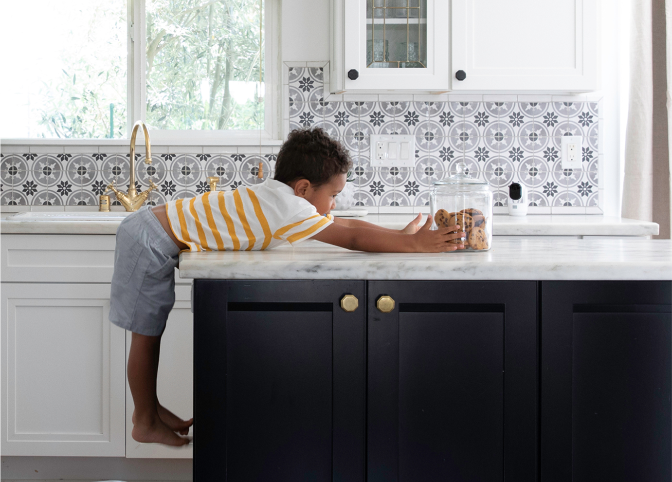 A boy reaching for a jar of cookies on a counter with the Arlo Essential Pan Tilt Indoor camera on the counter in the kitchen