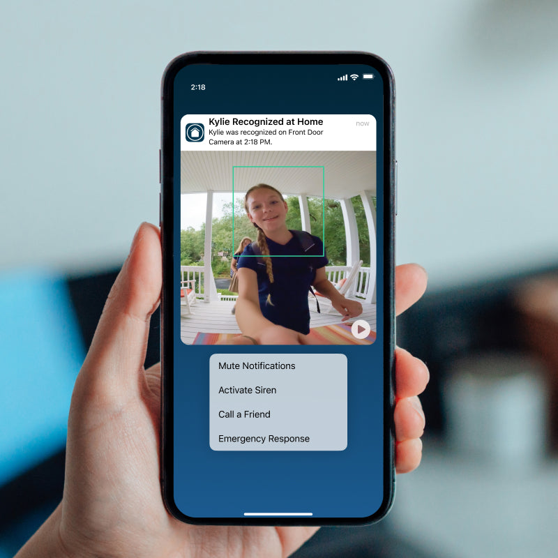 A hand holding a smartphone showing a Arlo Secure home security app notification on the screen, with a person visible in the on-screen video feed and a blurred indoor background.