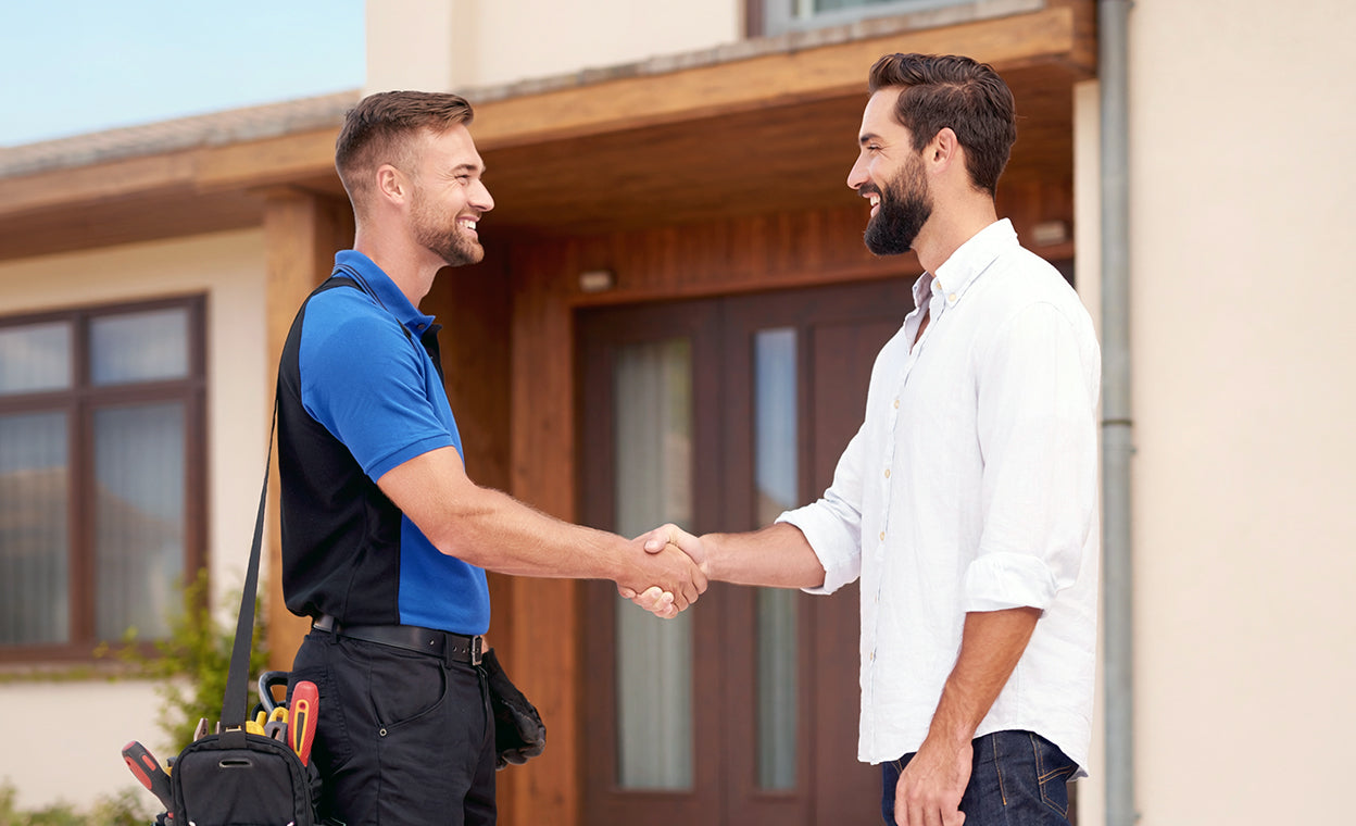 Two men shaking hands in front of a house, one with tools, indicating a professional interaction.