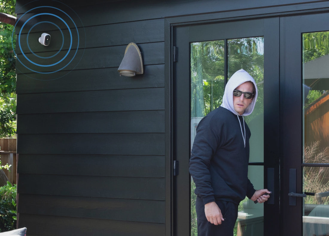 A burglar trying to open a door of a house and the Arlo Essential 3rd Gen XL camera siren pulsating on the side of a house